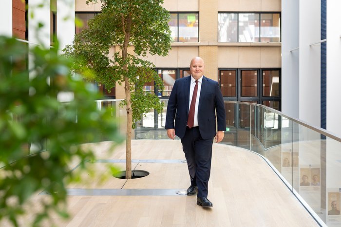 Man in donker pak met stropdas loopt over een glazen brug in een lichte kantoorhal met boom