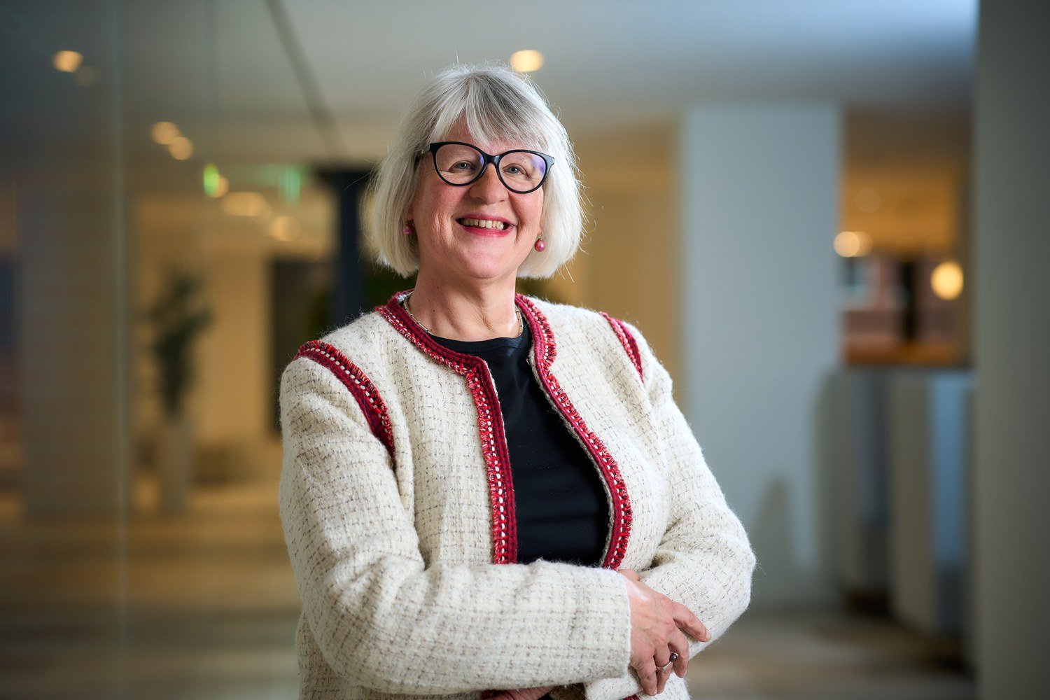 Vrouw met grijs haar en bril in wit met rood vest glimlacht in een lichte kantoorhal