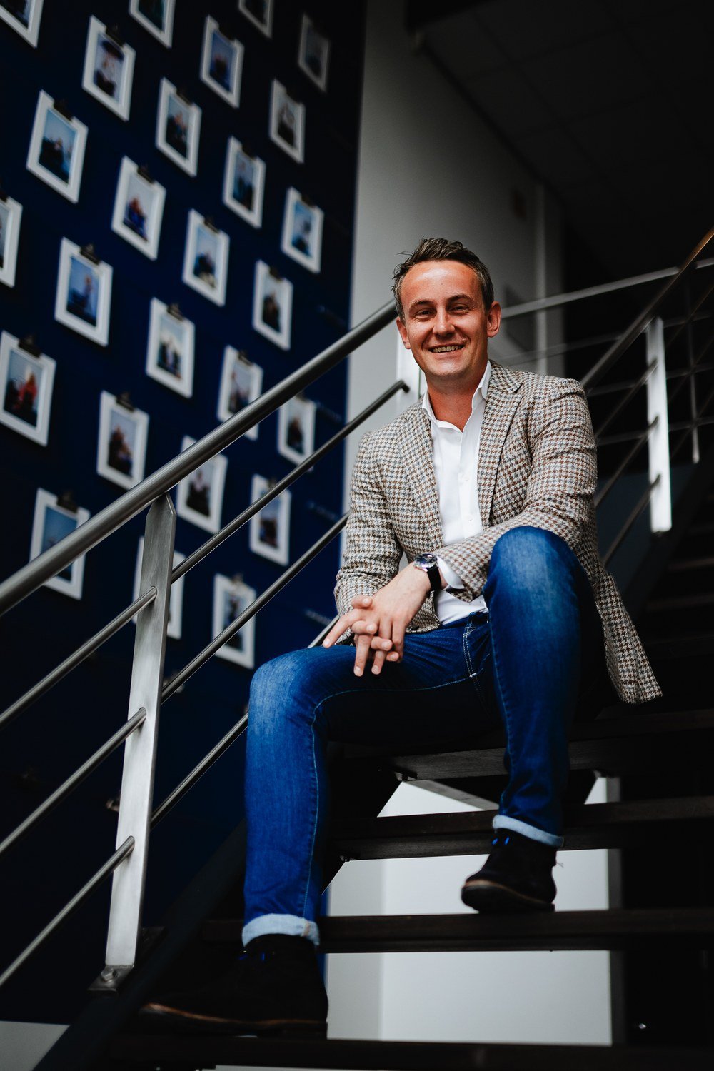 Lachende man in geruit colbert en spijkerbroek zit op een trap bij een muur met ingelijste foto's