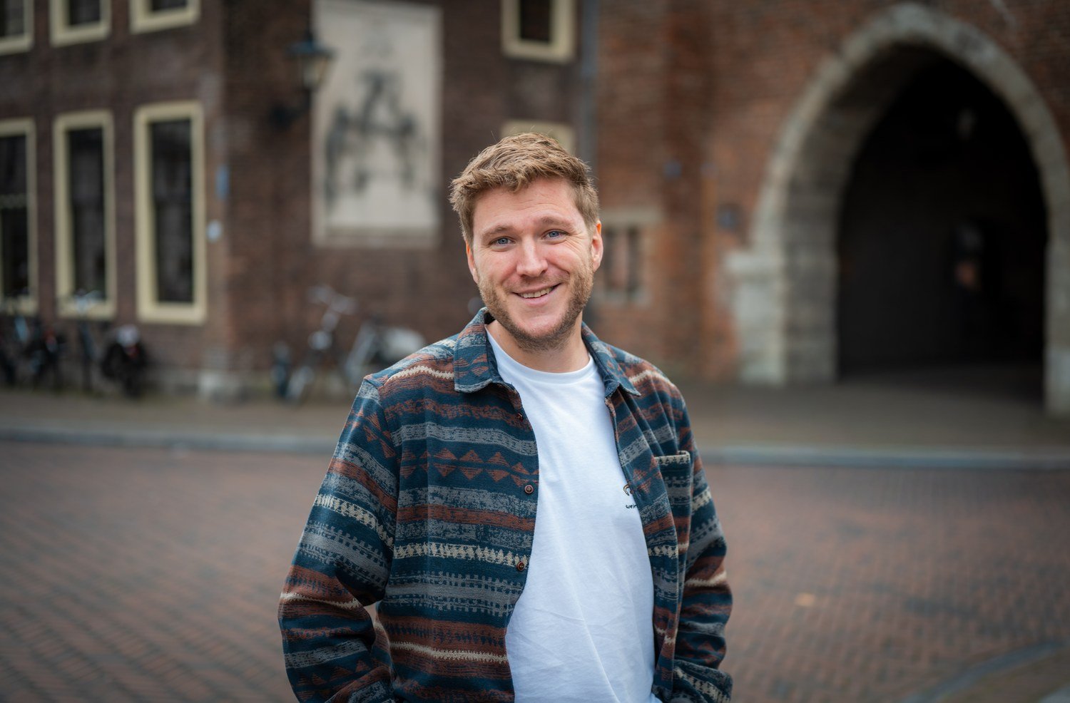 Lachende man met blond haar in gestreepte jas staat op een plein bij een oud bakstenen gebouw met poort