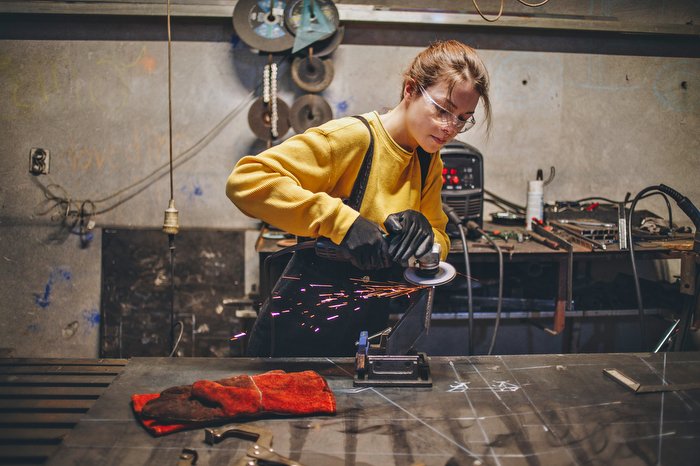 Vrouw met veiligheidsbril en werkhandschoenen slijpt metaal in een werkplaats, met vonken die alle kanten op spatten