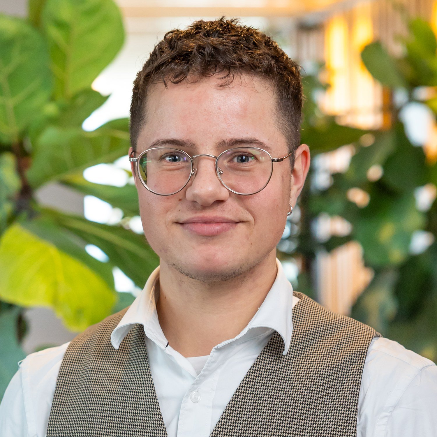 Portret van een jongeman met bril en gekruld haar, gekleed in een geruit gilet over een wit overhemd