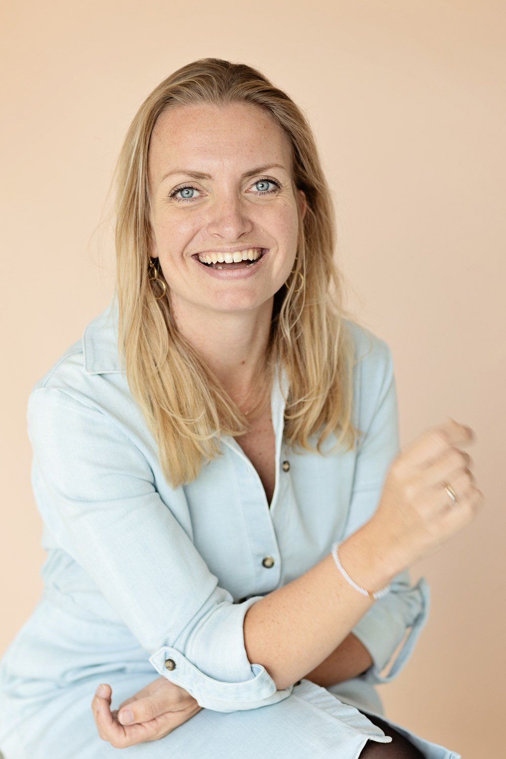 Lachende vrouw met blond haar in lichtblauwe blouse poseert tegen een beige achtergrond