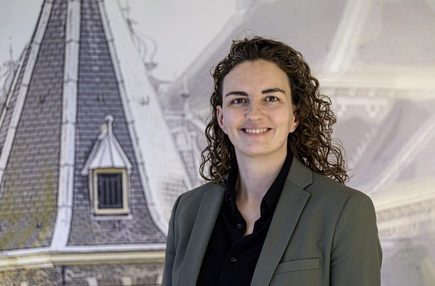 Lachende vrouw met krullend haar in blazer poseert voor een muurschildering van historische gebouwen met puntdaken